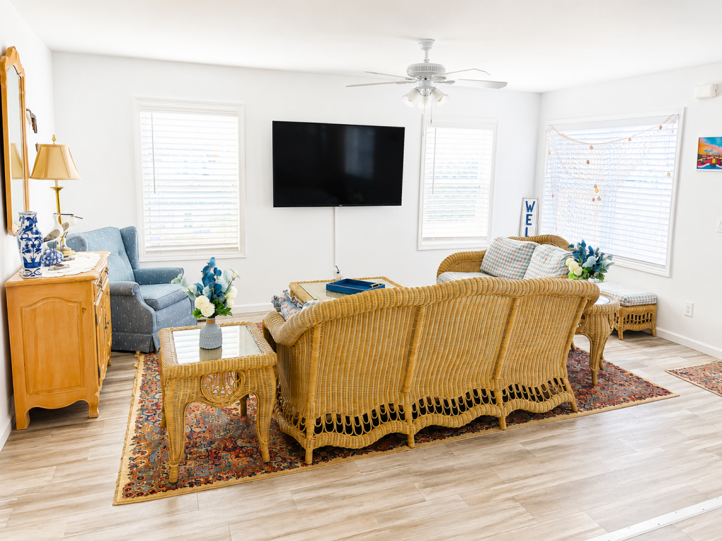 Bright, coastal-style living room at 212 E Rio Grande Avenue, Wildwood, NJ with wicker seating arranged around a glass-top coffee table, a large wall-mounted TV, blue accent decor, patterned area rugs and multiple windwos providing natural light