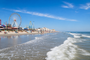 Wide sandy beach along the Jersey Shore with gentle ocean waves, blue skies and a bustling boardwalk featuring amusement rides in the distance on a sunny summer day.